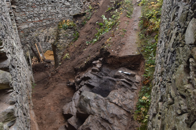 Snímek z jihozápadního koutu východního paláce na upravovaný skalní výchoz, kde se nacházelo přístupové schodiště. Výrazně černá vrstva ve střední části je pozůstatek požářiště s pozdně středověkými nálezy.  | © Autor F. Prekop